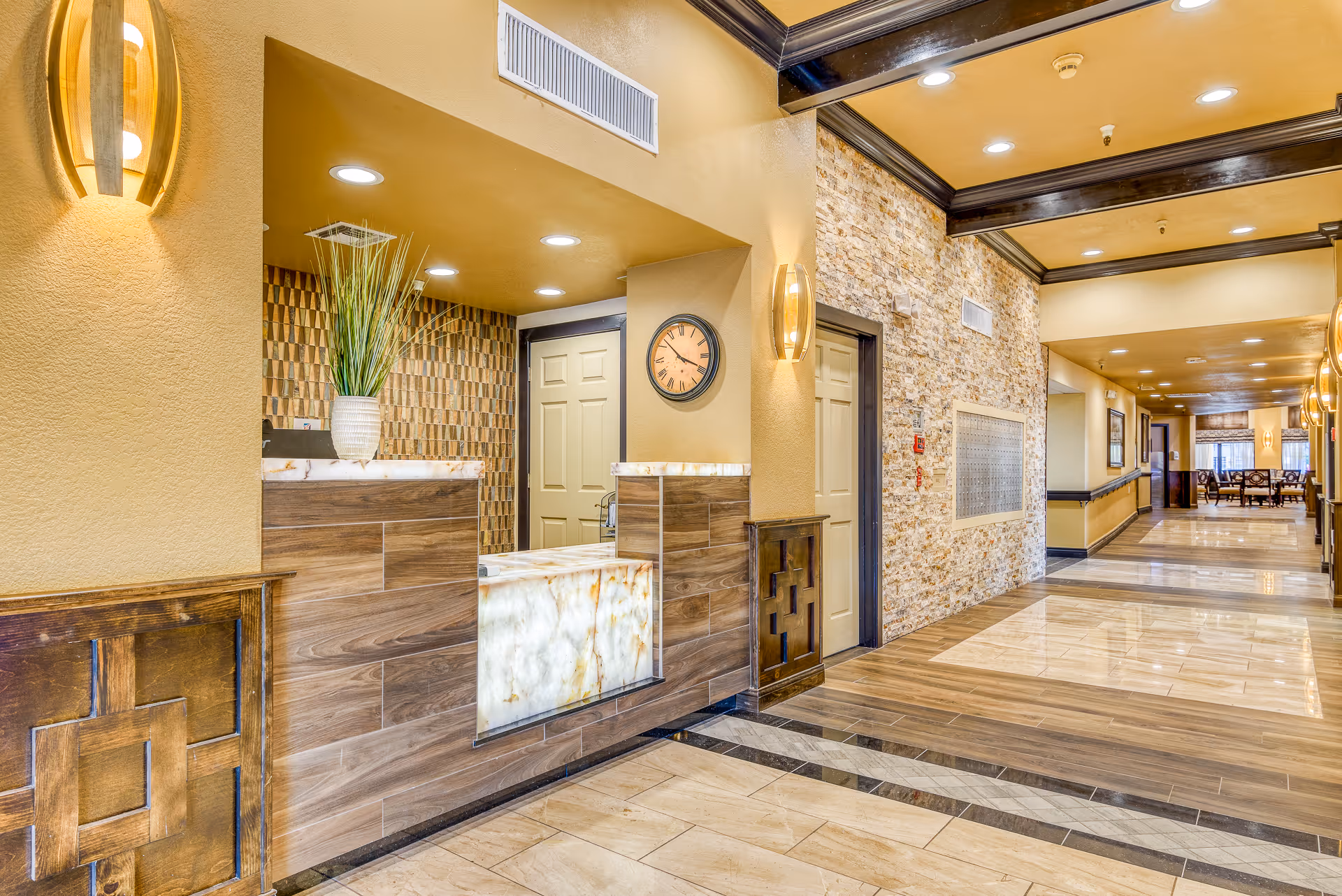 Interior view of a senior living facility hallway with a reception desk on the left, featuring a decorative plant and a wall clock. The hallway has warm yellow walls, recessed lighting, a stone accent wall, and a polished tile floor leading to a dining area with tables and chairs in the background.