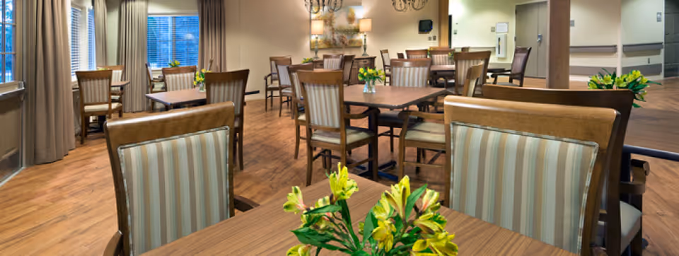 A well-lit dining room with multiple wooden tables and chairs featuring striped upholstery. Each table has a small vase with yellow flowers. Large windows with curtains allow natural light to enter the room, and the floor is made of wood. The walls are decorated with artwork and light fixtures.