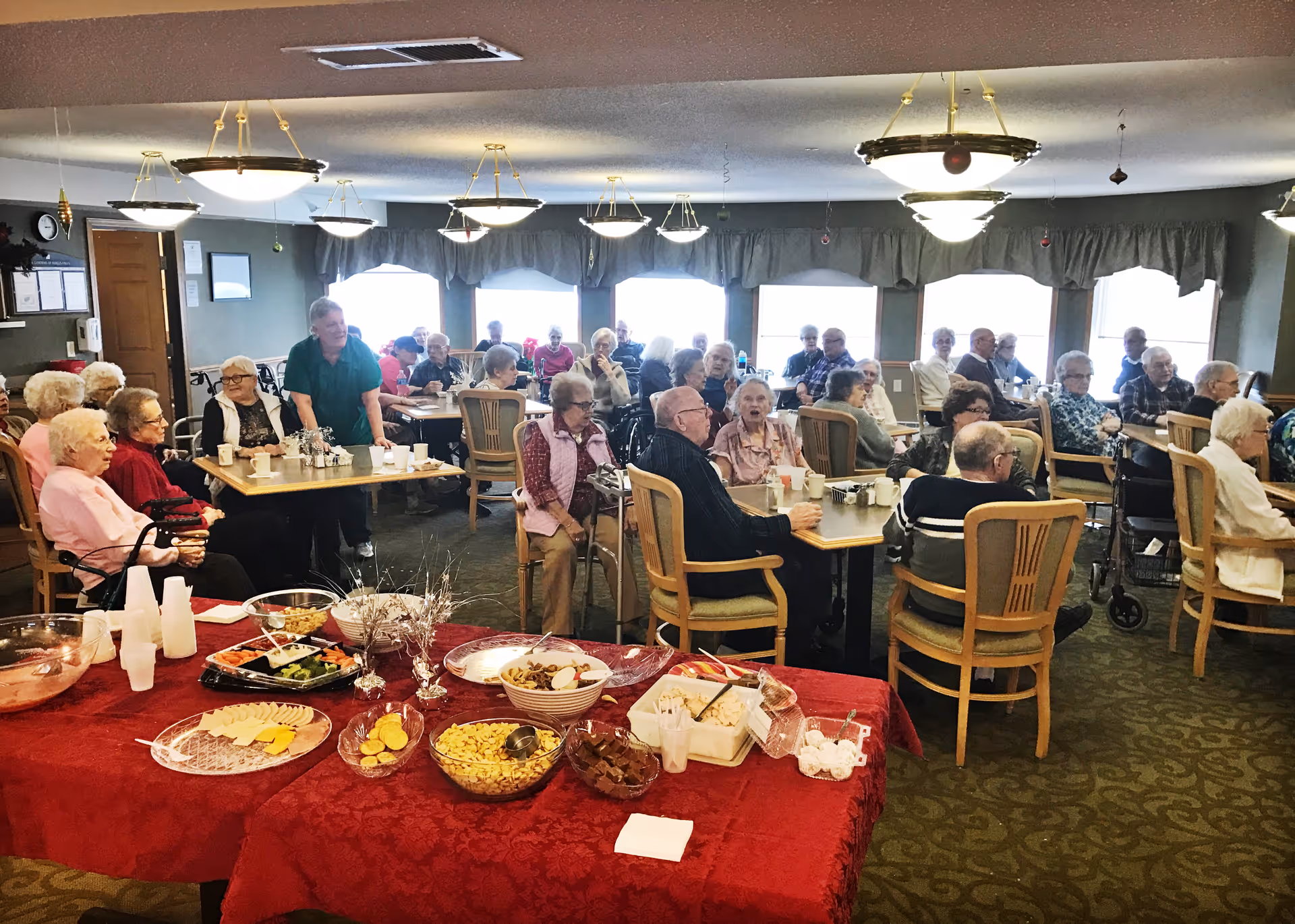A large group of elderly people seated at tables in a dining room setting, with a buffet table in the foreground covered with a red tablecloth and various snacks and dishes. The room is well-lit with multiple ceiling lights and large windows with curtains.