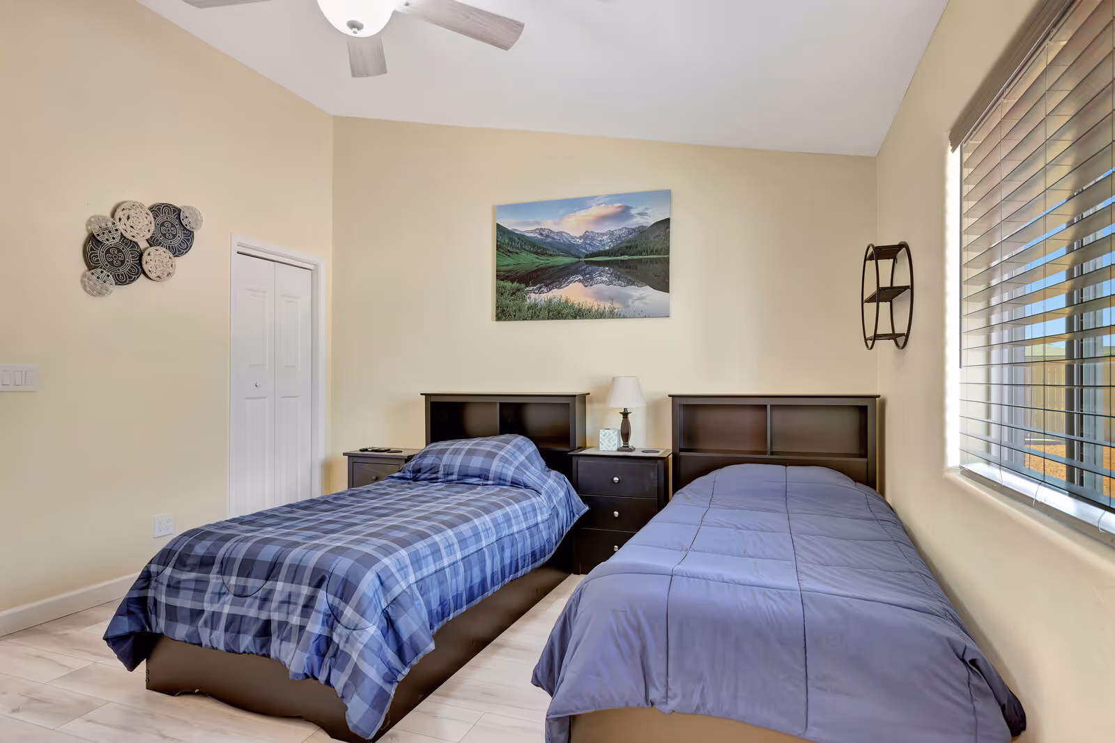 A bedroom with two single beds, one with a blue plaid comforter and the other with a solid blue comforter. Between the beds is a dark wooden nightstand with a lamp and a box of tissues. Above the nightstand is a wall-mounted picture of a mountain landscape with a lake reflecting the mountains. The room has beige walls, a ceiling fan, a window with blinds on the right, and decorative wall art on the left.