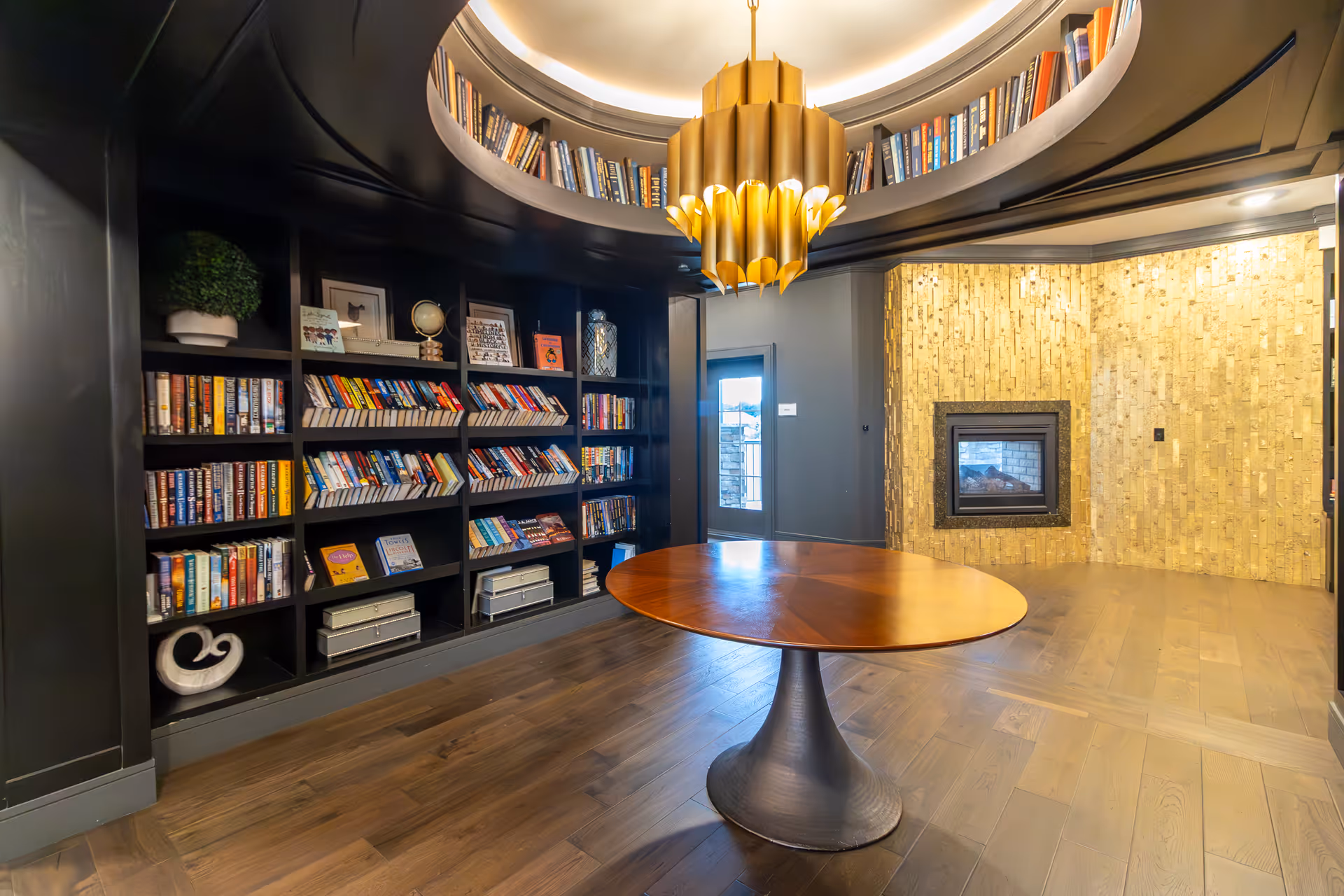 Modern common room with built-in bookshelves, round wooden table, gold chandelier and a fireplace set into a textured wall.
