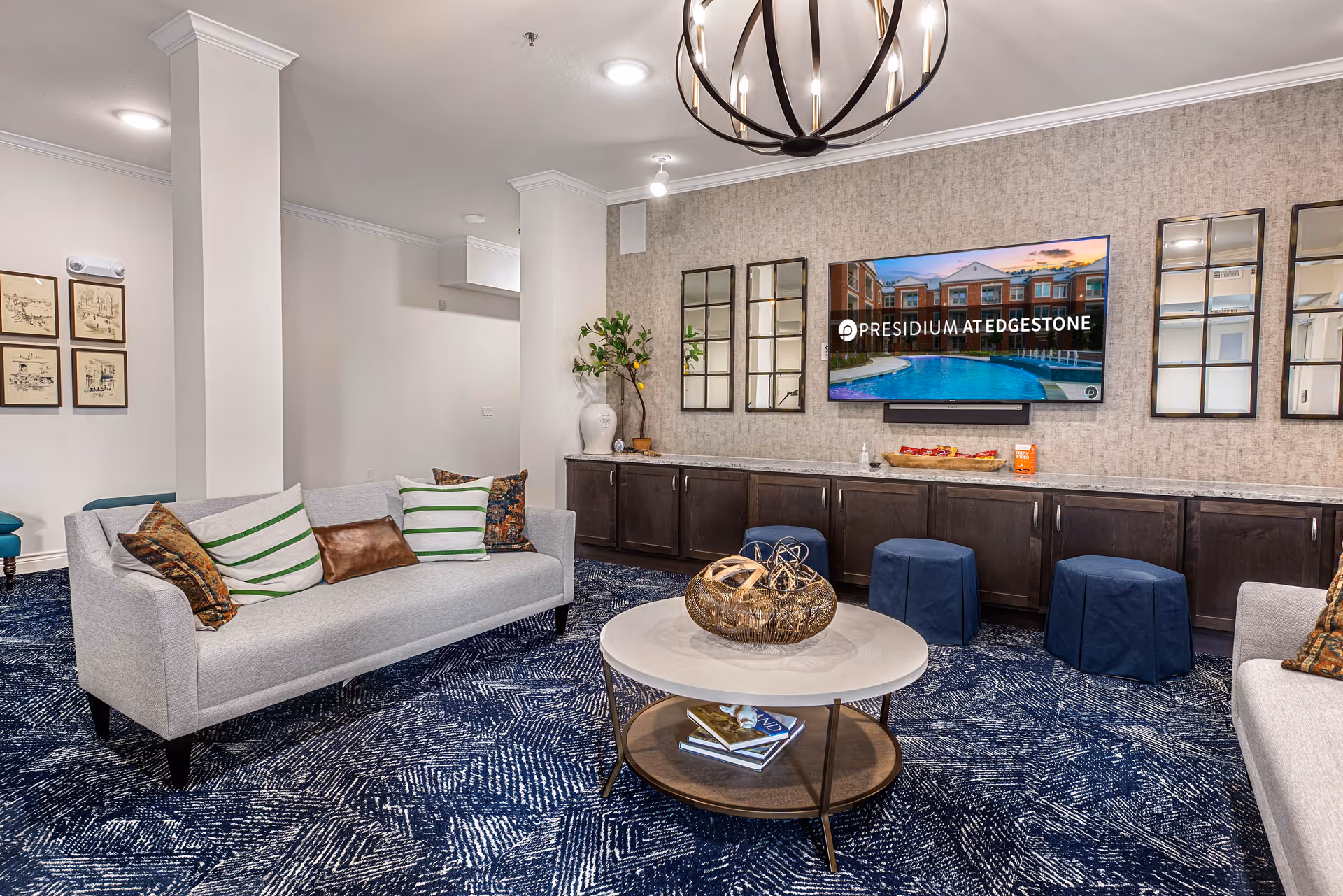 A modern living room area with a gray sofa adorned with decorative pillows, a round coffee table with decorative items and books underneath, and a wall-mounted TV displaying the Presidium at Edgestone logo and an image of a pool and building. The room features dark wood cabinetry, three blue stools, a patterned blue carpet, and a chandelier hanging from the ceiling. There are also framed pictures on one wall and three decorative mirrors on another.