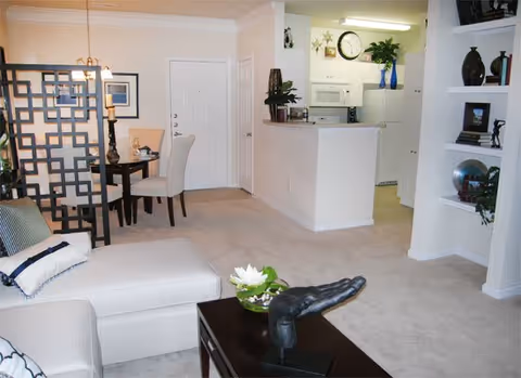 Interior view of a senior living facility showing a living room with a white chaise lounge and a dark coffee table with a decorative hand sculpture and a flower arrangement. In the background, there is a dining area with a small round table and two chairs, separated by a decorative room divider. The kitchen is visible beyond a half wall, featuring white cabinets, a microwave, and a clock on the wall. The walls are painted white, and the floor is carpeted in a light beige color.