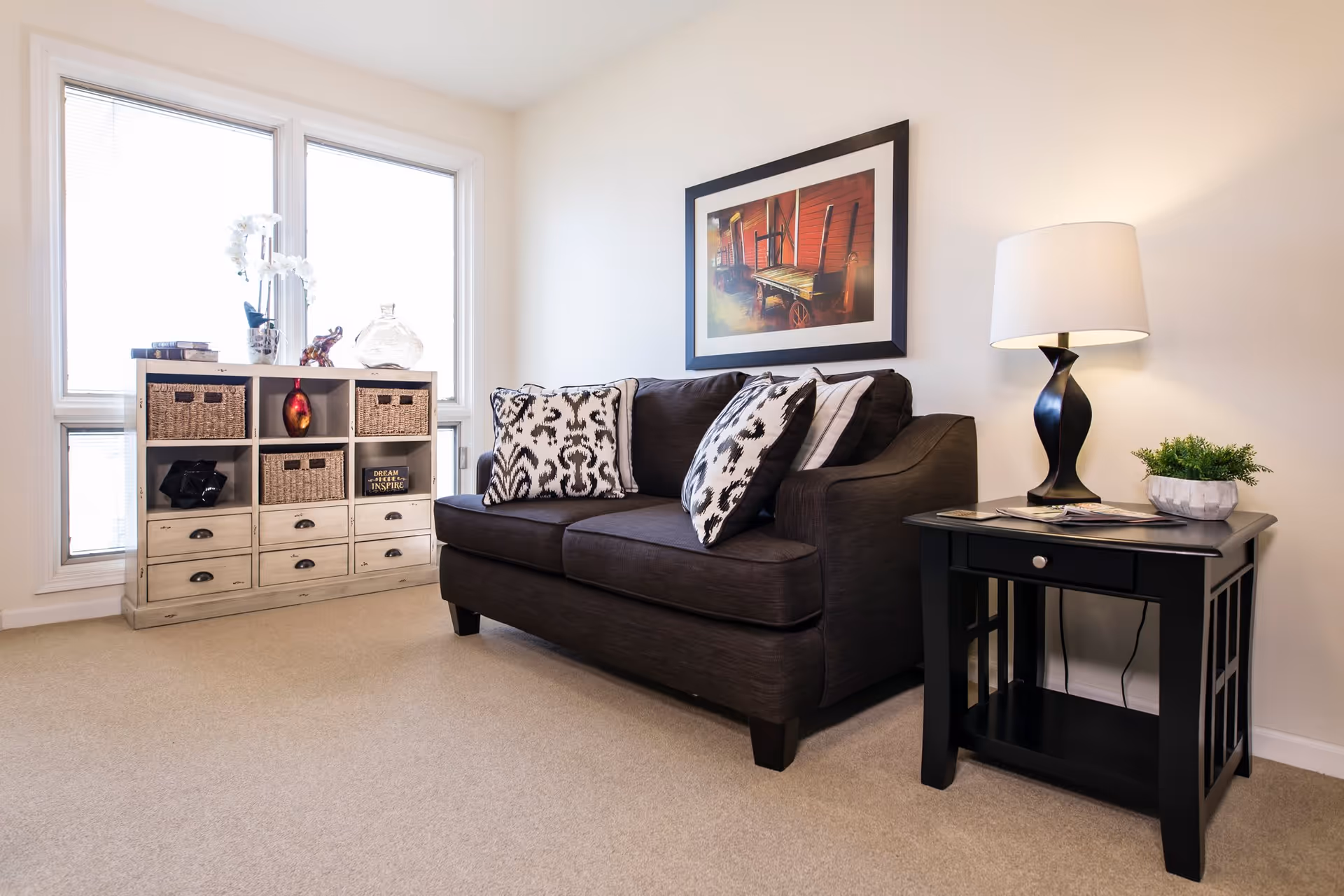 A cozy living room with a dark brown loveseat adorned with patterned pillows. To the right of the loveseat is a black side table with a modern black lamp and a small potted plant. On the wall above the loveseat hangs a framed picture of a rustic wagon. To the left, there is a white storage unit with baskets and decorative items, positioned in front of a large window letting in natural light. The room has beige carpeting and light-colored walls.
