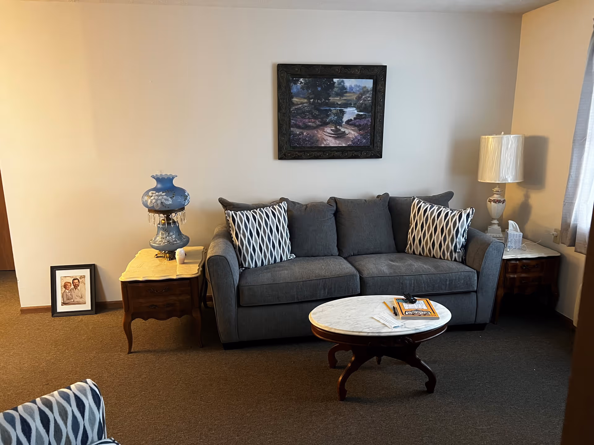 A cozy living room with a gray sofa adorned with two patterned pillows. In front of the sofa is a round wooden coffee table with a white marble top, holding a remote control, a notebook, a pen, and a face mask. On either side of the sofa are wooden side tables; the left one has a blue decorative lamp and a small white object, while the right one has a white lamp and a tissue box. A framed photo of a couple is placed on the floor leaning against the wall to the left. Above the sofa hangs a framed painting depicting a serene outdoor scene with trees and a river.