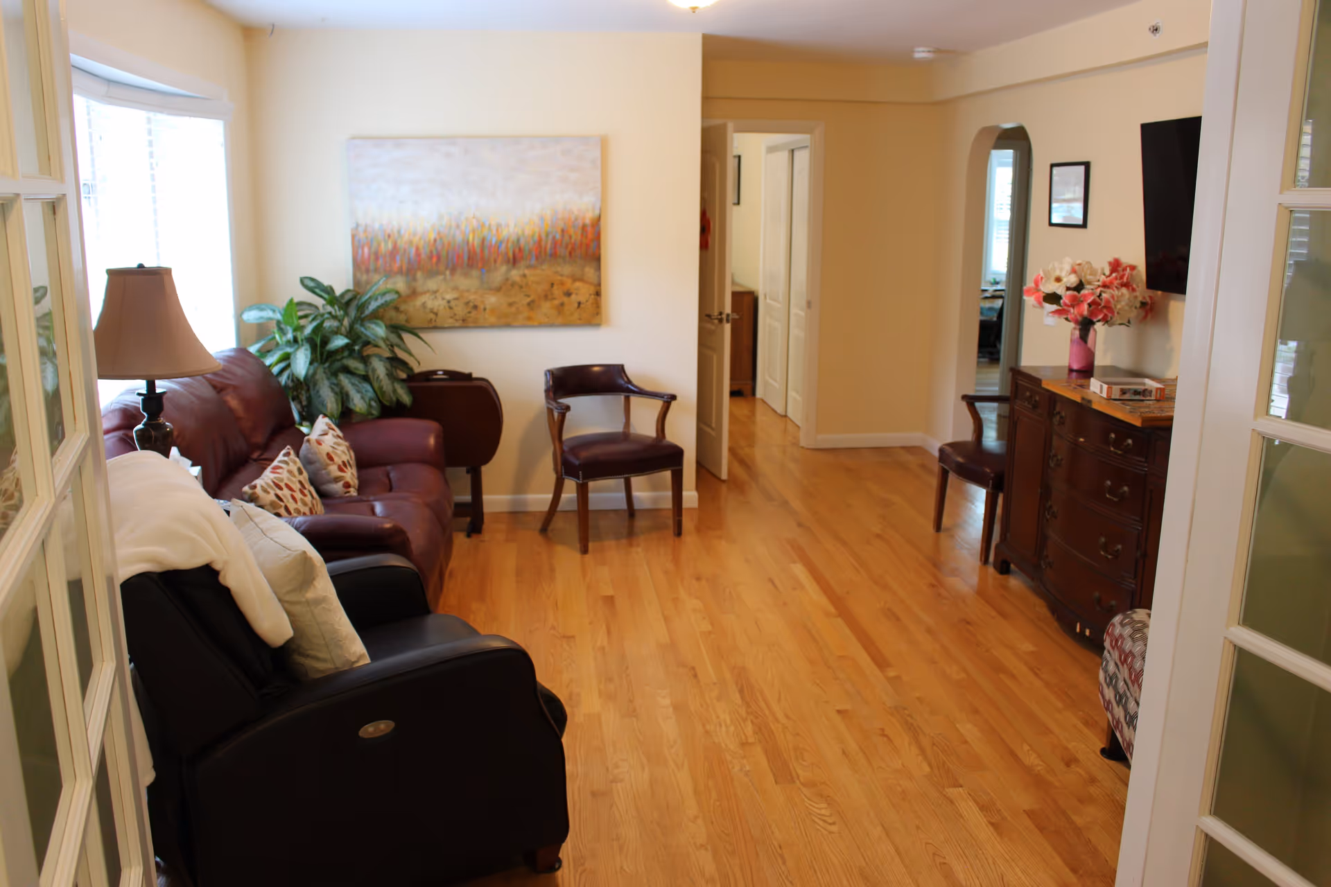 A cozy living room with wooden flooring, featuring a brown leather sofa with patterned cushions, a black recliner chair with a white blanket and pillow, a wooden armchair, a side table with a lamp, a large painting on the wall, a green potted plant, and a wooden dresser with a vase of flowers and a flat-screen TV mounted above it. The room is well-lit with natural light coming through a window with blinds.