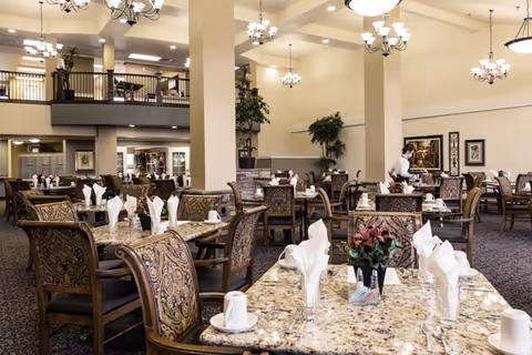 A spacious dining room with multiple tables set with white napkins, cups, and silverware. The room features high ceilings with chandeliers, patterned chairs, and decorative plants. A staff member is seen attending to one of the tables.