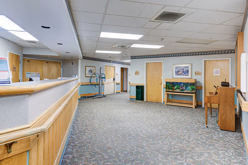 Interior hallway of a senior living facility with carpeted floor, light blue walls with wooden trim, several closed wooden doors, a fish tank on a wooden stand, a piano, and a reception desk on the left side.