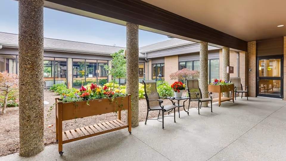 Covered outdoor patio area with stone columns, several chairs, small tables with flower pots, and raised flower beds. The patio overlooks a garden courtyard with various plants and shrubs, surrounded by a building with large windows.