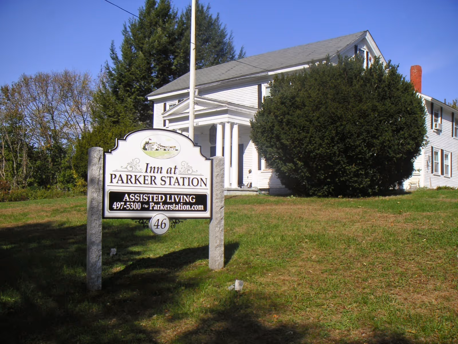 A roadside sign for "Inn at Parker Station" assisted living stands on the lawn in front of a white colonial-style house.