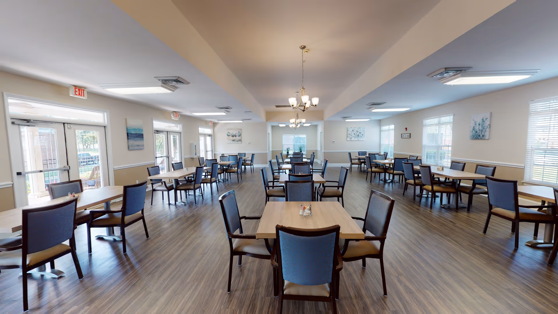 A spacious dining room with multiple wooden tables and blue cushioned chairs arranged neatly. The room has large windows allowing natural light to brighten the space, light fixtures hanging from the ceiling, and artwork on the walls. The floor is covered with wood-patterned flooring.