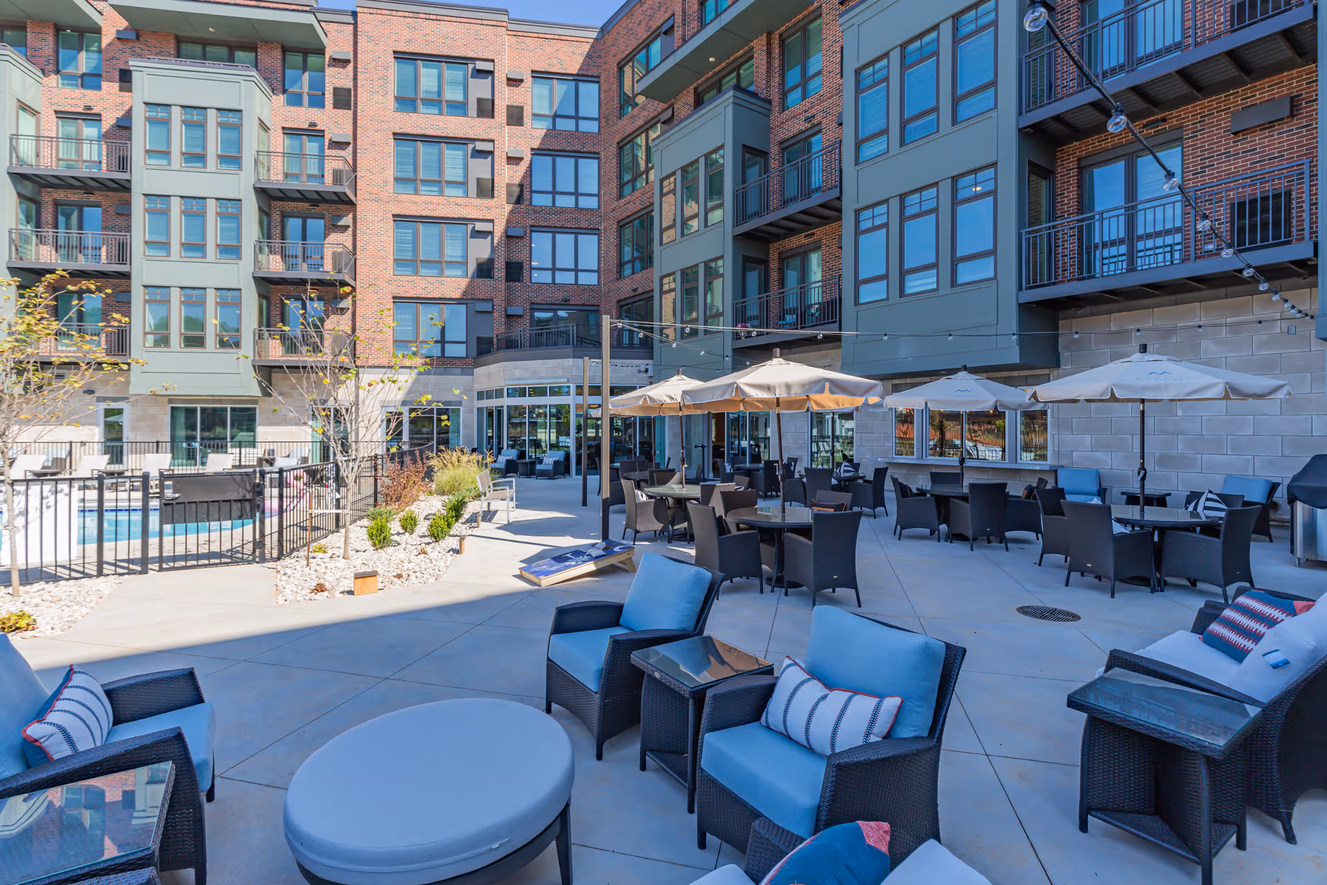 Outdoor patio area at Merrill Gardens at Greenville with multiple seating arrangements including cushioned chairs, tables with umbrellas, and a small pool area surrounded by a fence. The patio is adjacent to a multi-story brick and green-paneled building with many windows and balconies.