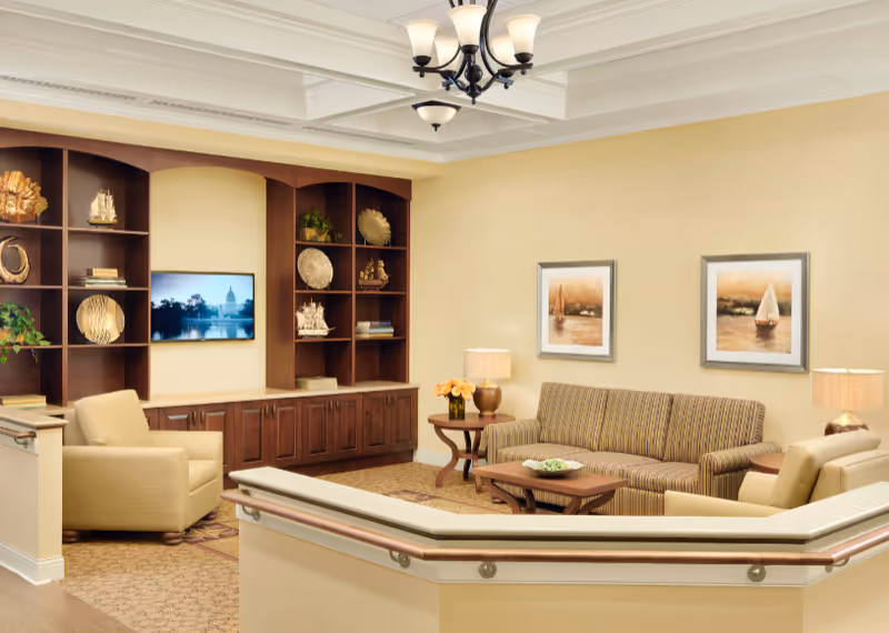 A cozy living room area with a striped sofa, two beige armchairs, a wooden coffee table, and a side table with a lamp and flowers. The walls are decorated with two framed paintings of sailboats. A built-in wooden shelving unit holds decorative items and a flat-screen TV. The ceiling features a chandelier light fixture.