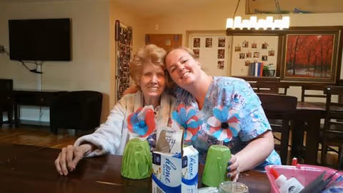An elderly woman and a caregiver sitting closely together at a wooden table, smiling at the camera. The table has craft items including colorful paper flowers and green paper cups. The background shows a living room area with a TV mounted on the wall, a chair, and framed pictures on the wall.