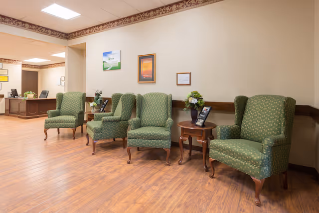 A waiting area with four green upholstered armchairs arranged along a beige wall with a wooden chair rail. Between the chairs are small wooden tables holding flower arrangements and informational brochures. The floor is wood laminate, and the ceiling has recessed lighting. In the background, there is a reception desk with office chairs and framed pictures on the walls.