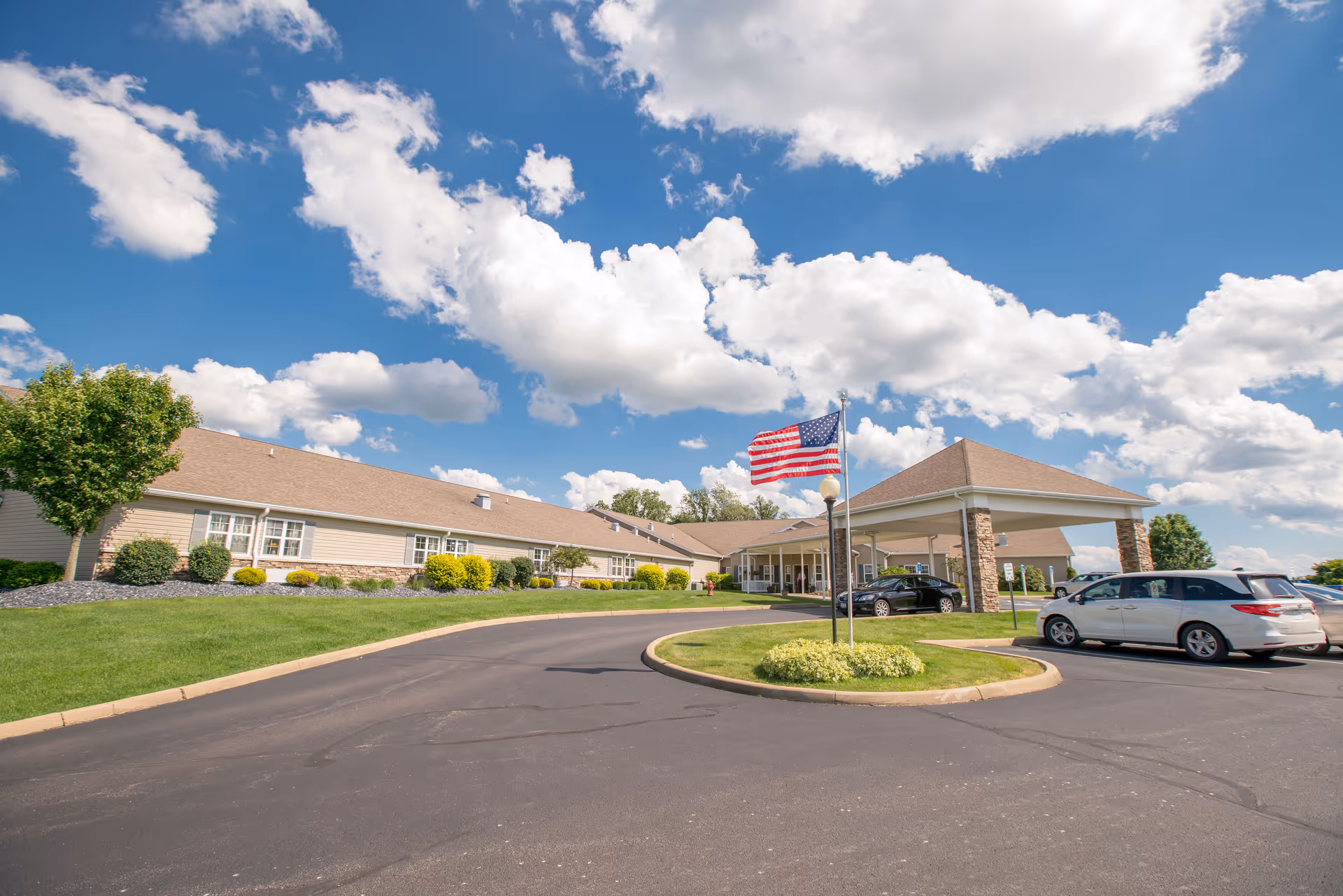 Exterior view of Cumberland Pointe Care Center building under a blue sky with scattered clouds. The building has a beige facade with stone accents and a covered entrance. An American flag is flying on a flagpole in front of the entrance. Several cars are parked in the parking lot nearby, and there is well-maintained landscaping with green grass and bushes.