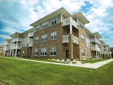 Exterior view of a three-story senior apartment building with brick and light-colored siding, multiple balconies, and a well-maintained lawn under a partly cloudy sky.