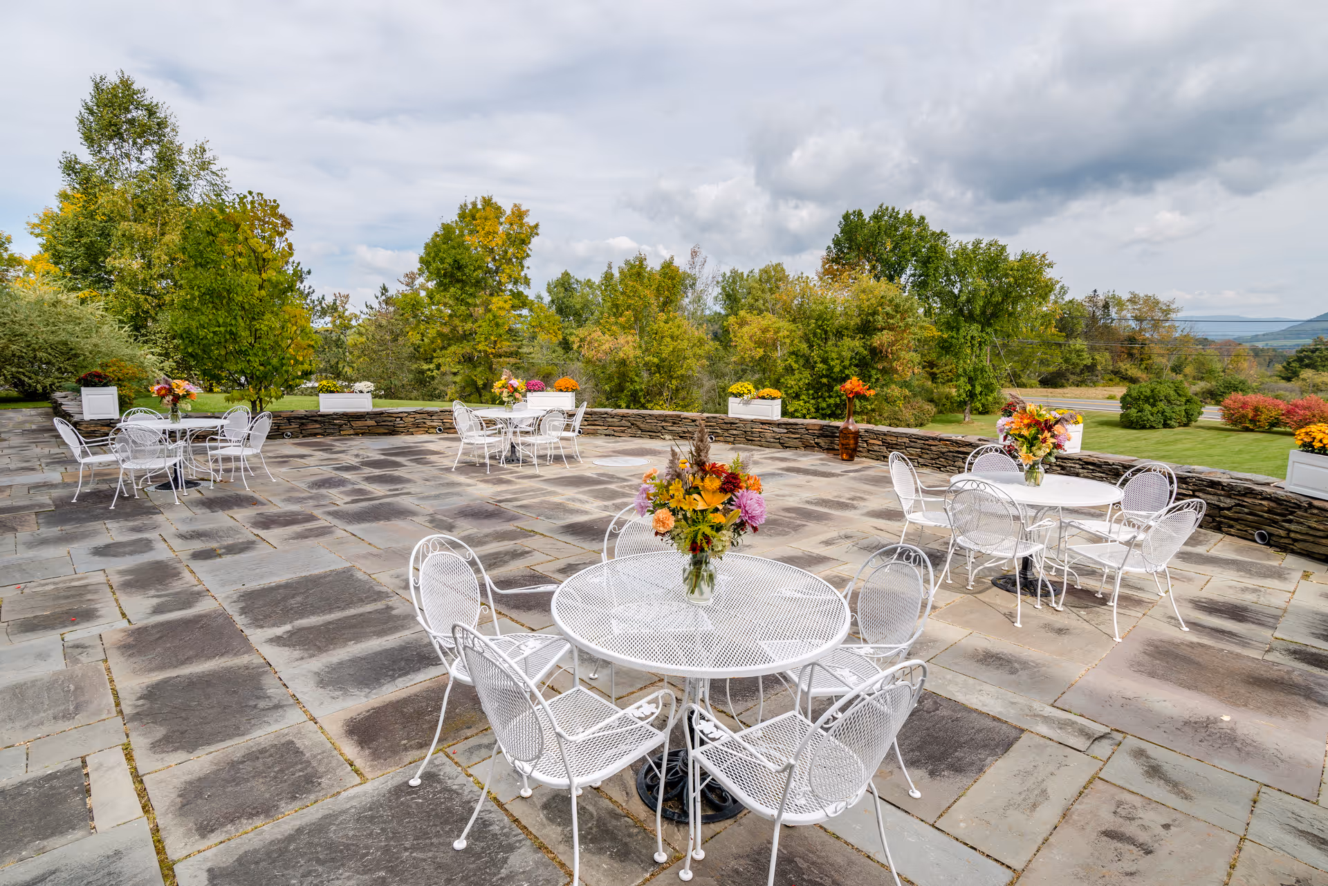 Stone patio with white metal tables and chairs arranged with floral centerpieces, overlooking trees and distant hills.
