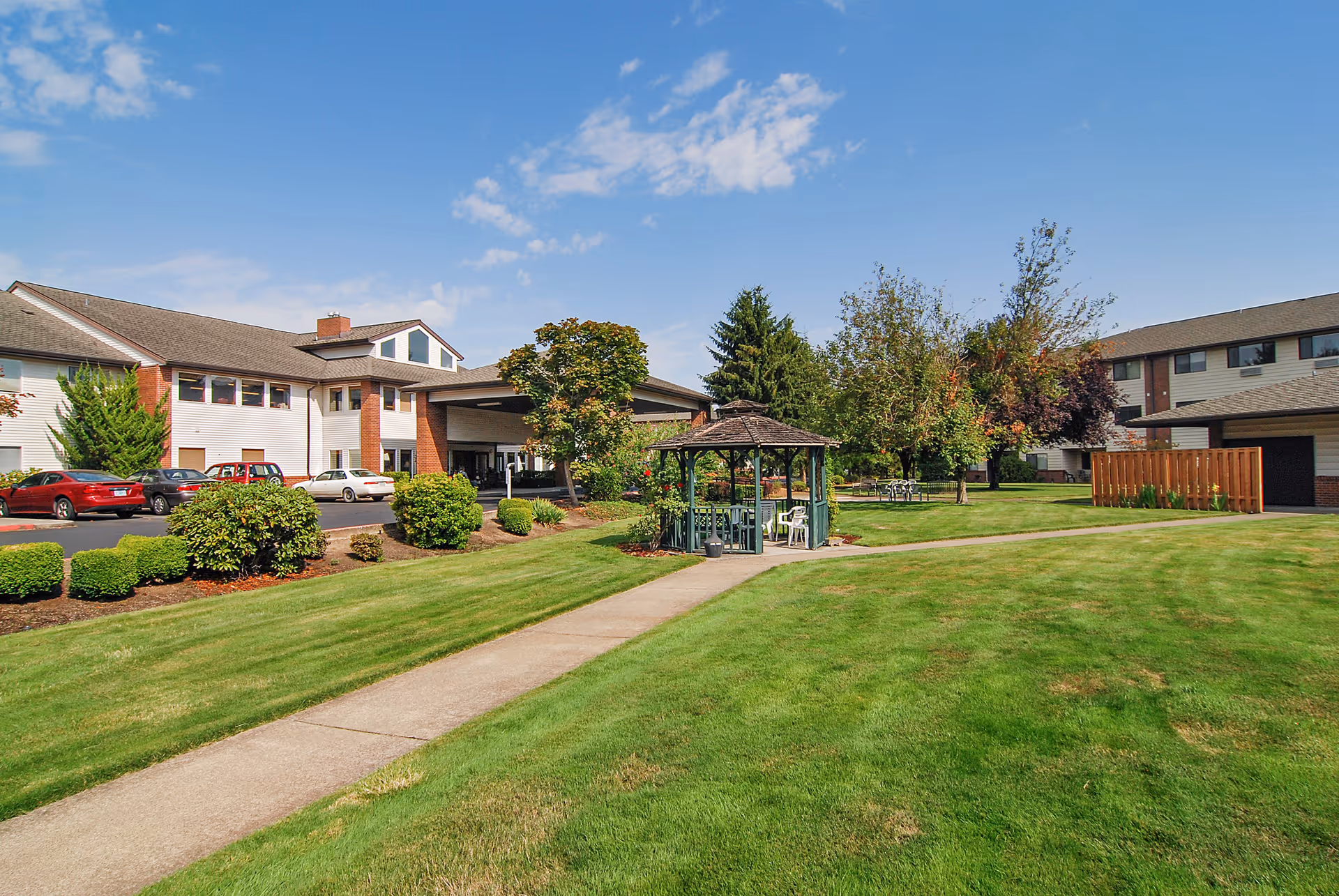 Exterior view of Brookdale McMinnville City Center showing a well-maintained lawn with a concrete walkway leading to a green gazebo surrounded by trees and shrubs. The building is a two-story structure with white siding and brick accents, and several parked cars are visible near the entrance under a covered drop-off area. The sky is clear with a few clouds.