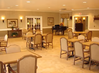 Spacious dining/activity room with multiple tables and chairs, a grand piano and warm lighting.