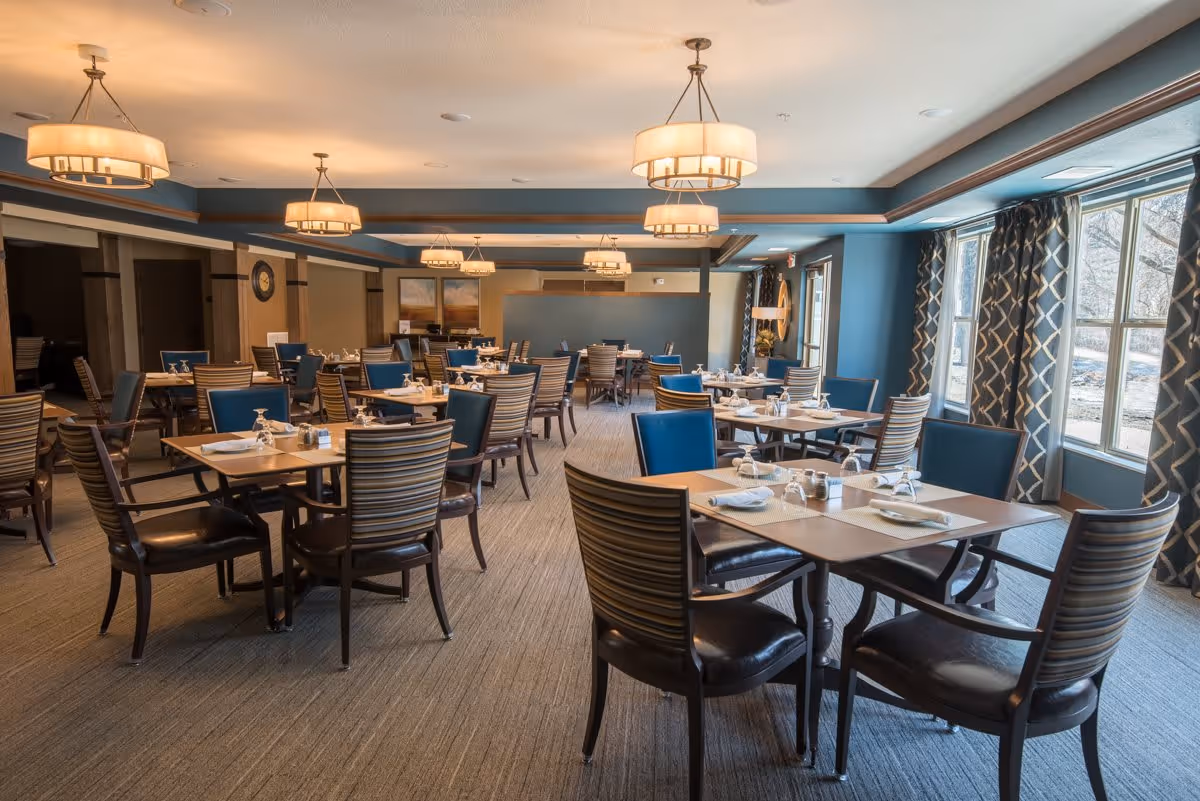 A spacious dining room with multiple wooden tables and chairs arranged neatly. Each table is set with napkins, glasses, and salt and pepper shakers. The room features large windows with patterned curtains allowing natural light to fill the space. Modern ceiling lights hang from the ceiling, and the walls are painted in a combination of blue and beige tones.