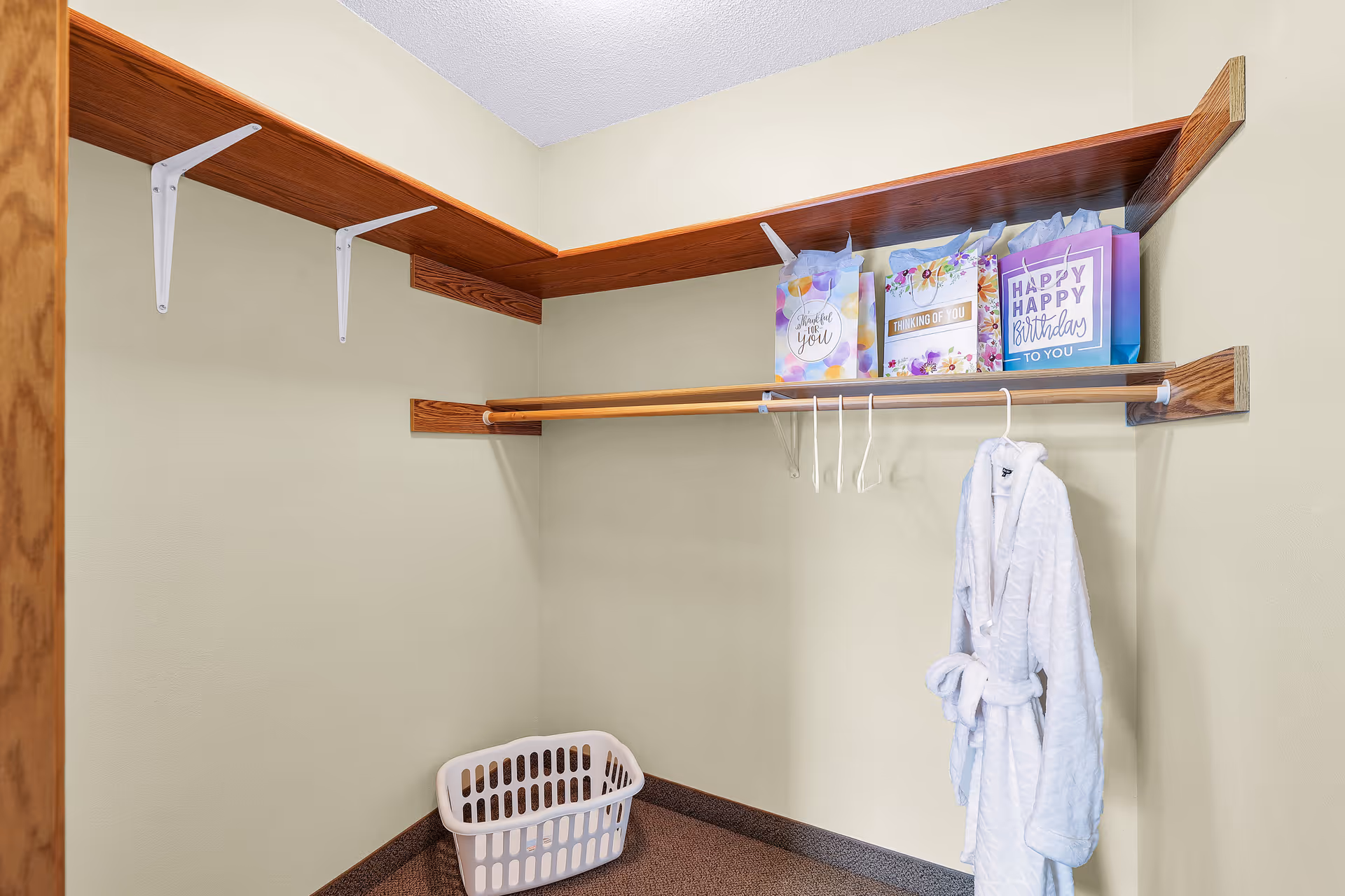 A small walk-in closet with wooden shelves and a hanging rod. There are three colorful gift bags on the top shelf, a white bathrobe hanging on a hanger, and a white laundry basket on the floor.