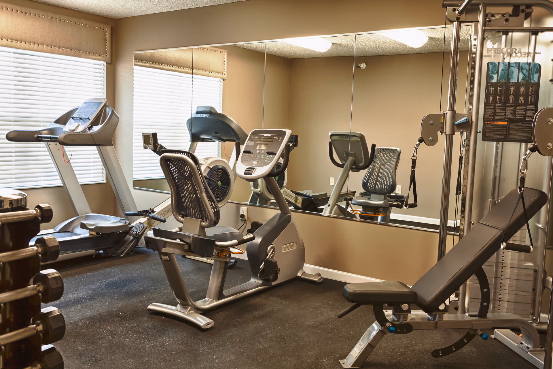 A fitness room with exercise equipment including a treadmill, a recumbent stationary bike, a rowing machine, a weight bench, and a multi-functional weight machine. There is a large mirror on the wall reflecting the equipment and two windows with blinds letting in natural light.