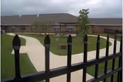 Wrought-iron fence in the foreground overlooking a landscaped courtyard with winding concrete paths and a single-story brick nursing facility under a cloudy sky.