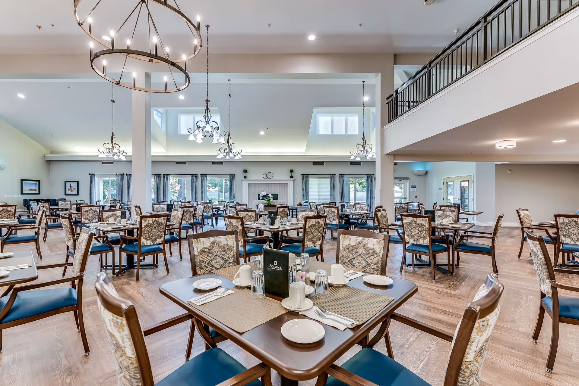 Spacious dining room in a senior living facility with multiple tables and chairs arranged neatly. Each table is set with plates, cups, napkins, and utensils. The room features large windows with curtains, elegant chandeliers hanging from the ceiling, and a high ceiling with an upper balcony area visible.