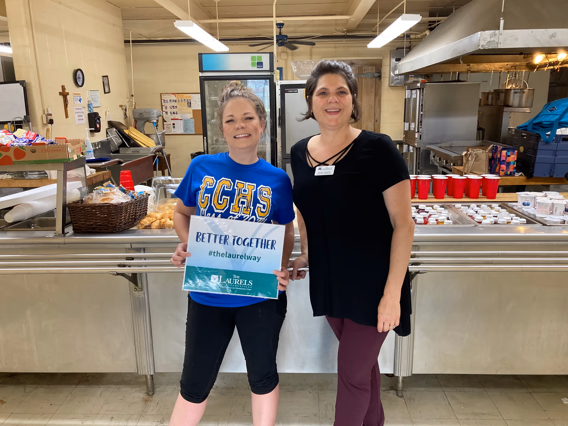 Two women standing in a kitchen area. One woman is wearing a blue CCHS shirt and holding a sign that says 'BETTER TOGETHER #thelaurelway The Laurels of Steubenville'. The other woman is wearing a black top and maroon pants with a name tag. Behind them are kitchen counters with food items, red cups, and kitchen equipment.