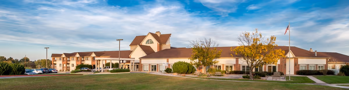 Wide exterior view of Emporia Presbyterian Manor, a large single-story building with a pitched roof, surrounded by a well-maintained lawn, trees with autumn foliage, benches, a flagpole with an American flag, and a parking area with cars under a partly cloudy sky.