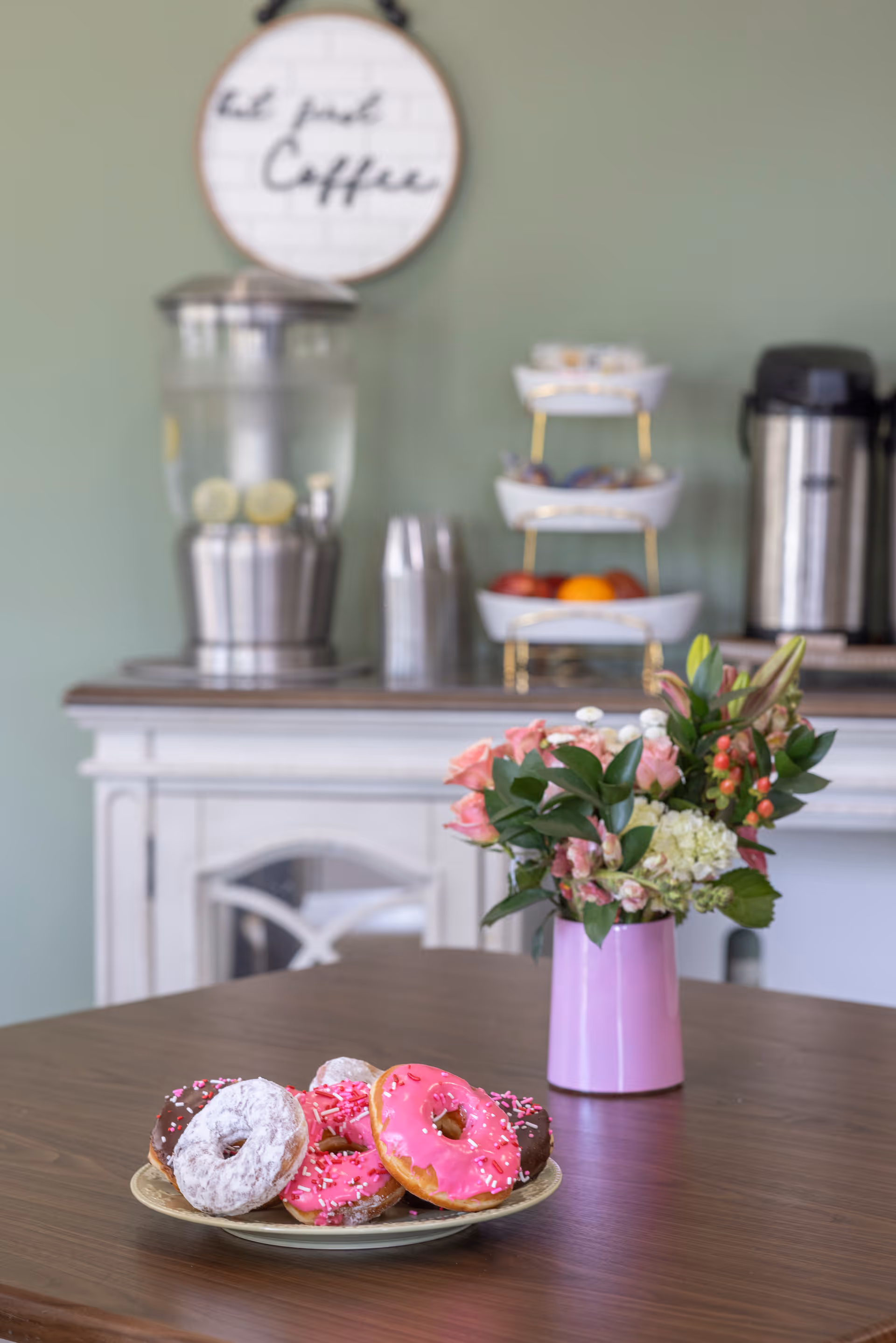 A plate of pink-iced and powdered donuts and a vase of flowers on a wooden table with a beverage station (water dispenser, coffee urn, and fruit tiers) in the background.