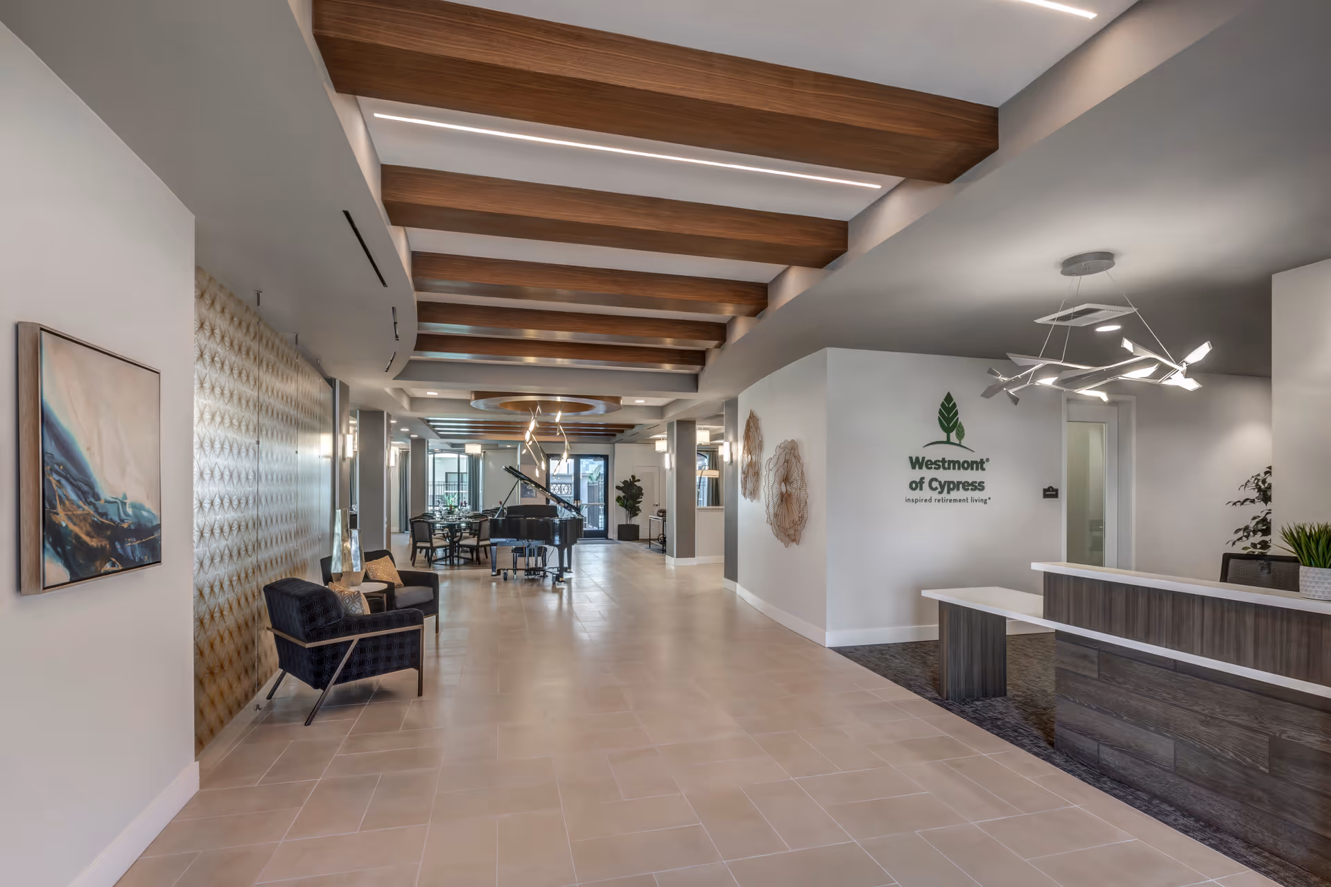 Spacious and modern interior lobby area of Westmont of Cypress featuring a reception desk on the right, decorative wall art, a seating area with chairs on the left, a grand piano in the background, and wooden ceiling beams with contemporary lighting fixtures.