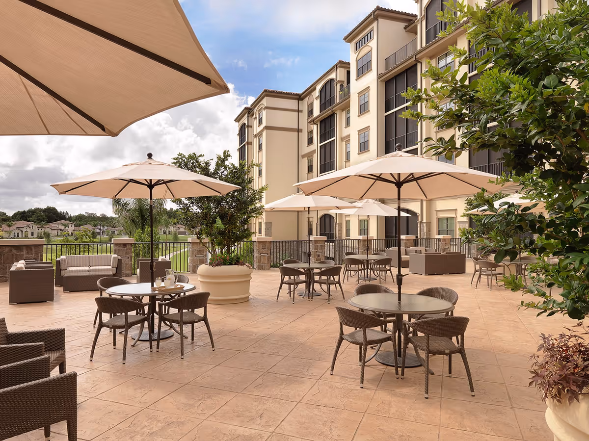 Outdoor patio area at The Arlington of Naples featuring multiple round tables with chairs and large beige umbrellas. The patio is spacious with tiled flooring, potted plants, and comfortable seating arrangements. The building exterior is visible in the background under a partly cloudy sky.