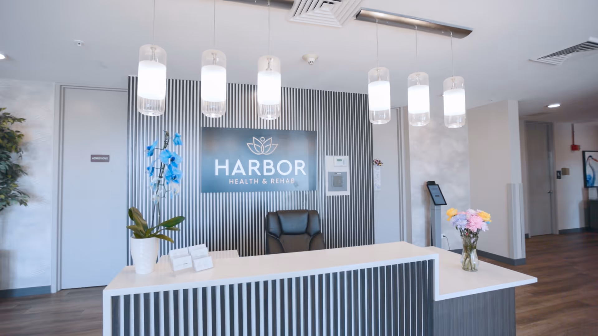 Reception desk area inside Harbor Health & Rehab facility with a modern design. The desk has a white countertop with vertical black and white striped paneling. There are two flower arrangements on the desk, one with blue flowers and one with mixed pastel flowers. Behind the desk is a black and white striped wall with a sign that reads 'HARBOR HEALTH & REHAB'. Above the desk hang six cylindrical pendant lights. The floor is wood, and there are doors and a plant visible in the background.