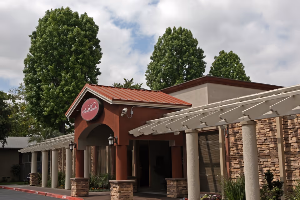 Exterior view of Southland Care Center featuring a covered entrance with stone pillars and a red roof. The building has a stone facade and is surrounded by tall green trees under a partly cloudy sky.