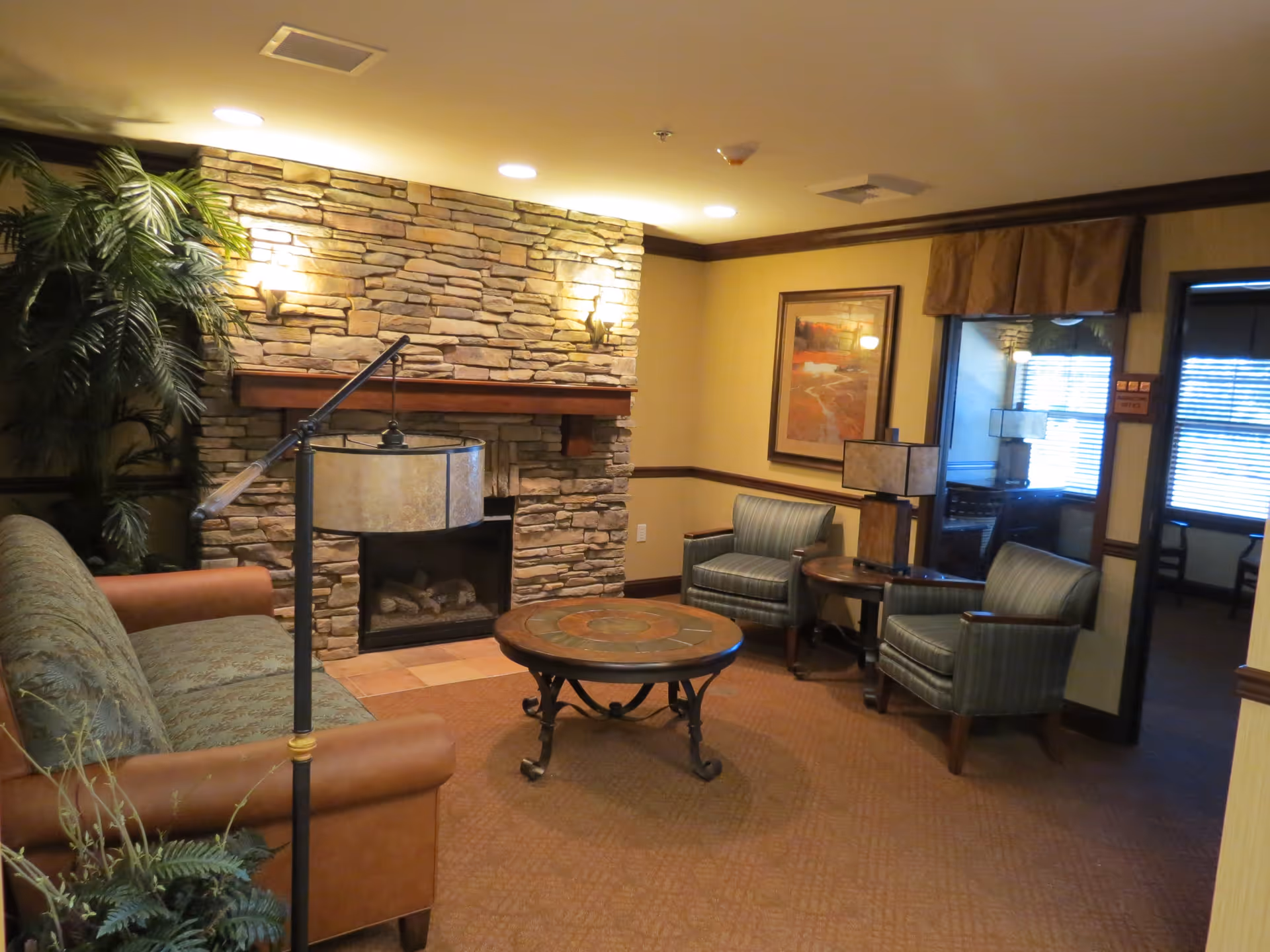 A cozy living room area with a stone fireplace, a brown leather and fabric sofa, two striped armchairs, a round coffee table, a side table with a lamp, framed artwork on the wall, and a large plant in the corner. The room has warm lighting and carpeted flooring.