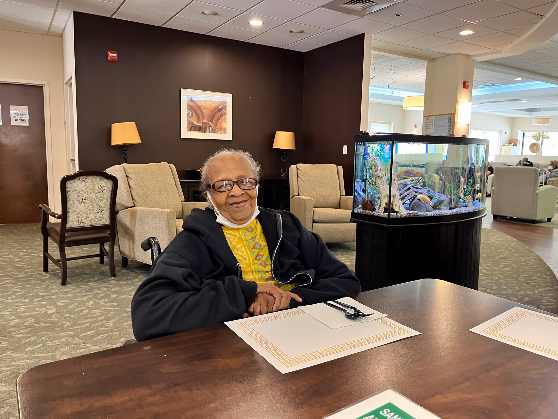 An elderly woman wearing glasses and a black jacket sits at a wooden table with placemats and utensils in a comfortable lounge area. Behind her are beige armchairs, a dark brown accent wall with a framed picture, two lamps, and a large fish tank. The room has a carpeted floor and a bright, open layout with additional seating in the background.