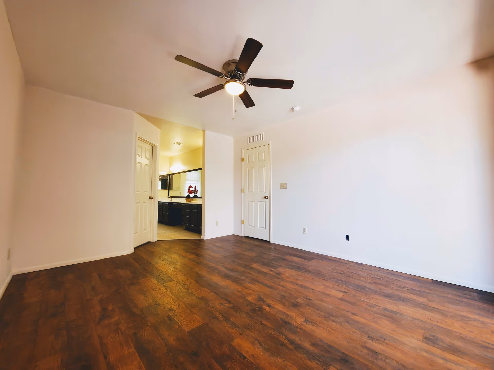 Empty room with wooden flooring, white walls, a ceiling fan with light, and two closed doors. One door leads to a bathroom area with a large mirror and dark cabinetry.