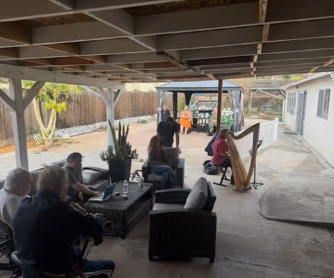 Covered outdoor patio area with several elderly people sitting on wicker furniture and a person playing a harp. In the background, there is a gazebo and a fenced yard with trees and plants.