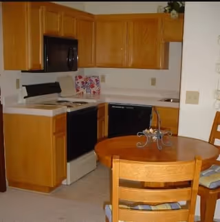 A small kitchen area with wooden cabinets, a black microwave, a black dishwasher, a white stove, and a round wooden dining table with two chairs. There is a decorative item on the table and a colorful item on the kitchen counter.