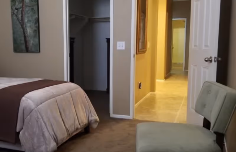 A bedroom with a bed covered in a beige comforter and a brown throw blanket. There is a green cushioned chair in the foreground and a hallway with beige walls and tiled floor visible through an open door. A closet with a dark interior is also visible.