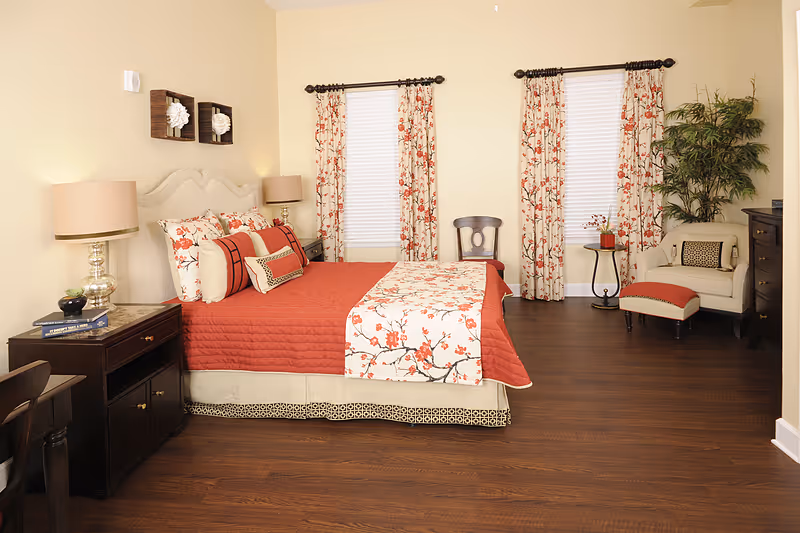 A well-decorated bedroom with a large bed featuring a red quilt and floral-patterned pillows and runner. The room has two windows with matching floral curtains, a wooden nightstand with a lamp and books, a small chair, a round side table with a plant, an armchair with a matching ottoman, and a tall green plant in the corner. The floor is dark wood and the walls are painted light beige.