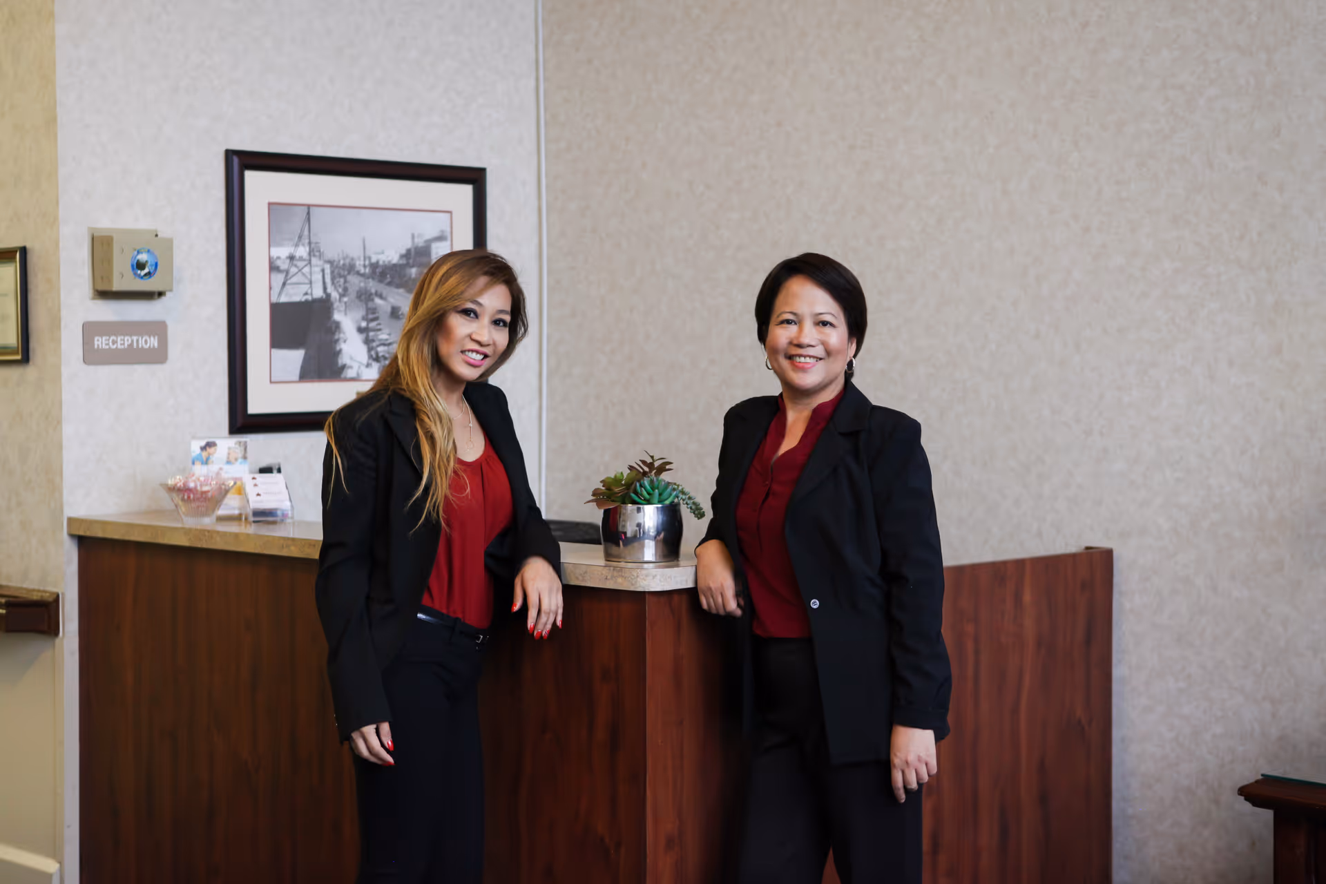 Two women standing and smiling behind a wooden reception desk in an indoor setting. One woman has long blonde hair and is wearing a black blazer over a red top, while the other has short dark hair and is also dressed in a black blazer with a red top. There is a small potted plant on the desk, a framed black and white photograph on the wall, and a sign labeled 'RECEPTION'.