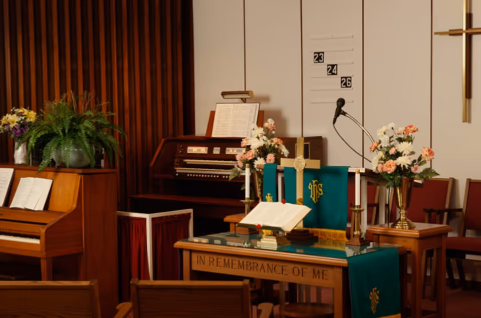 Interior of a chapel or worship area with a wooden altar draped in green cloth with gold religious symbols, an open Bible, candles, and flower arrangements. Behind the altar is a microphone, hymn board with numbers 23, 24, and 26, a wooden organ, and a piano with sheet music. Wooden chairs are visible in the foreground.