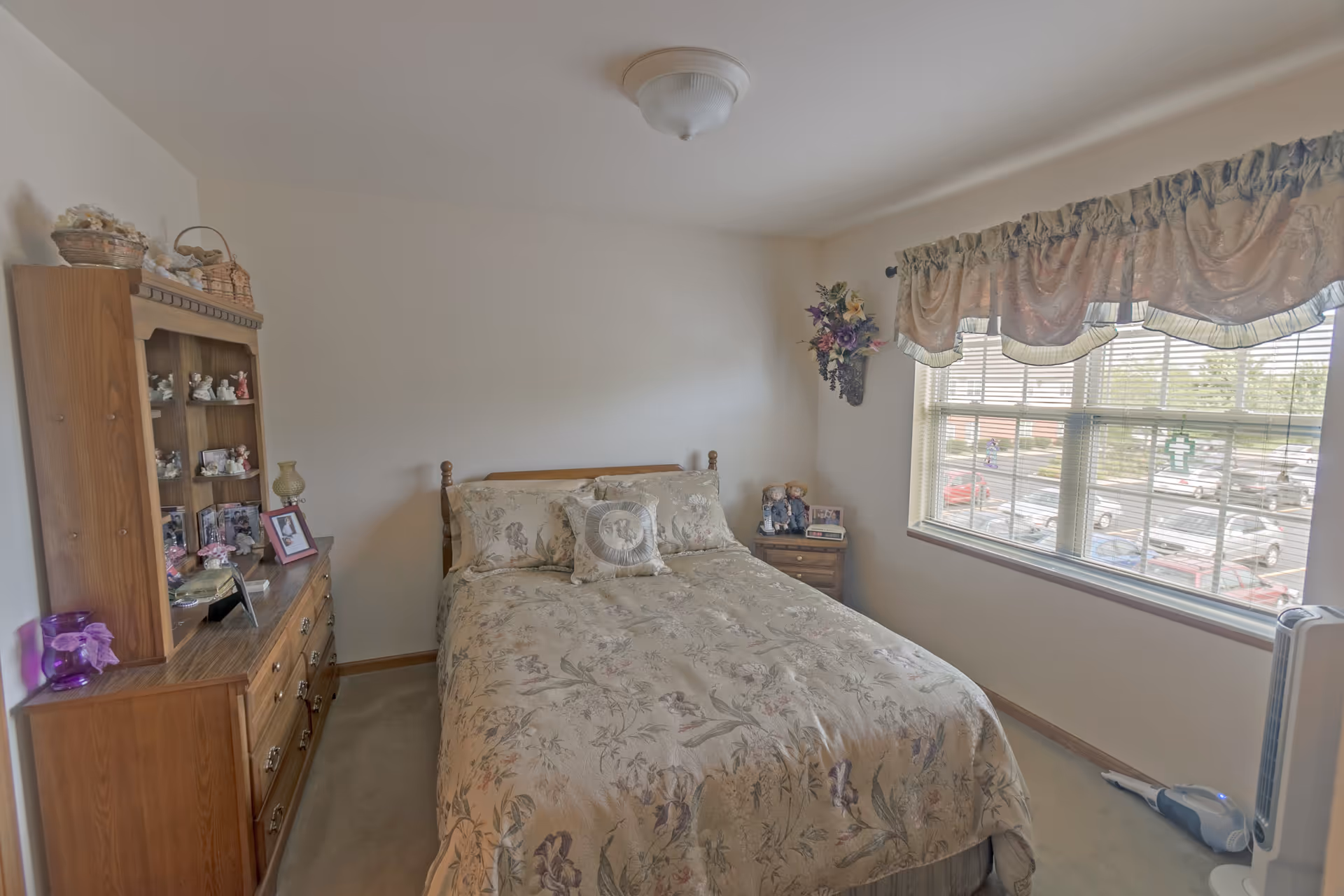 A cozy bedroom with a floral-patterned bedspread on a wooden bed. To the left is a wooden dresser with a hutch displaying small figurines and framed photos. A window with floral valance curtains lets in natural light, showing a parking lot outside. A small nightstand with decorative dolls and a clock is beside the bed, and a white fan and vacuum cleaner are on the floor near the window.