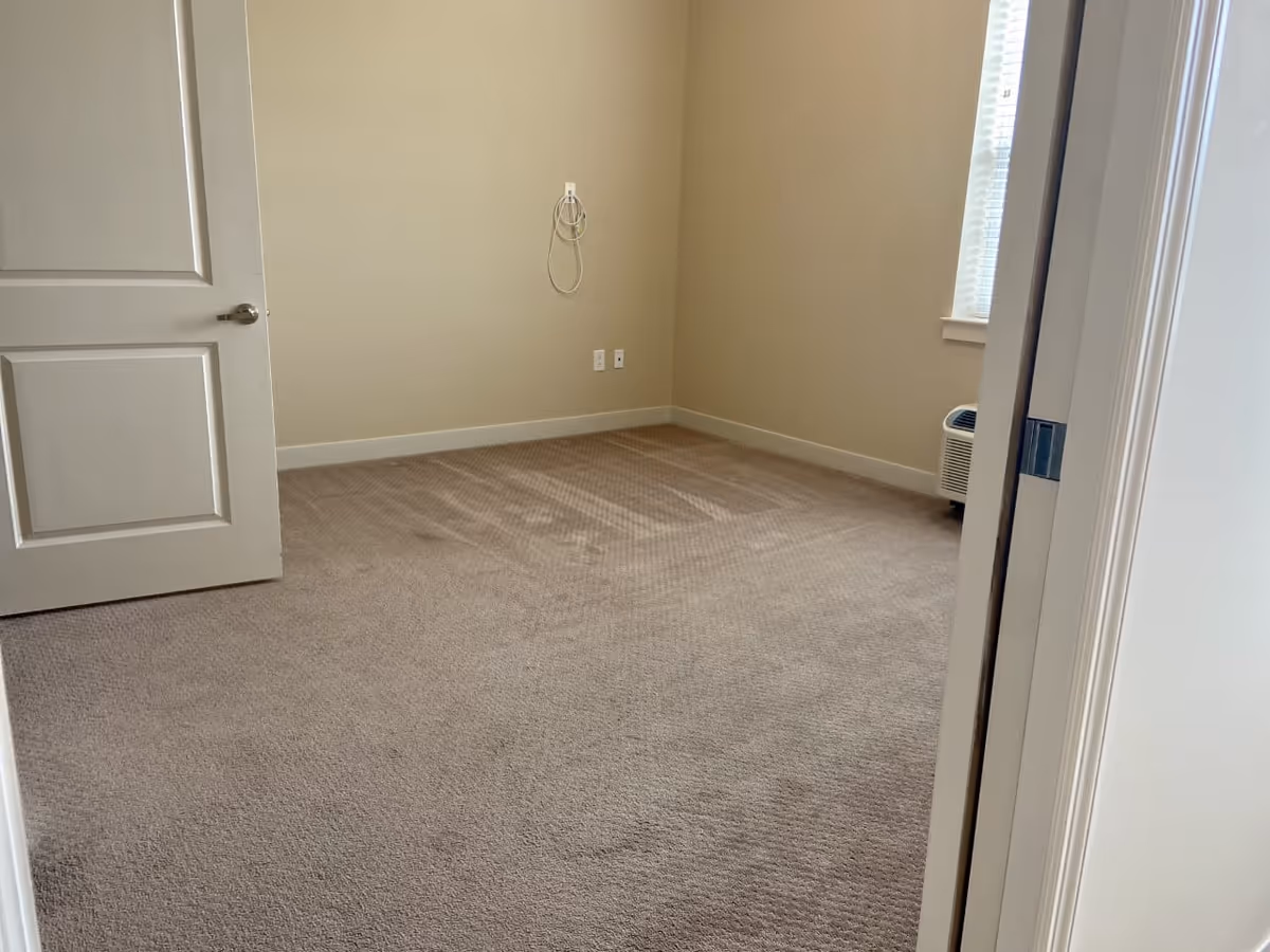 Empty carpeted bedroom with beige walls, an open door, a window and a portable air unit.