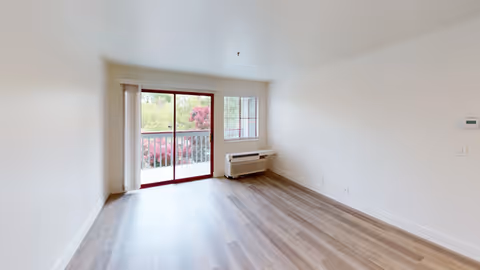 Empty room with light wood flooring, white walls, a large sliding glass door leading to a balcony with greenery outside, and a window with blinds next to a wall-mounted air conditioning unit.