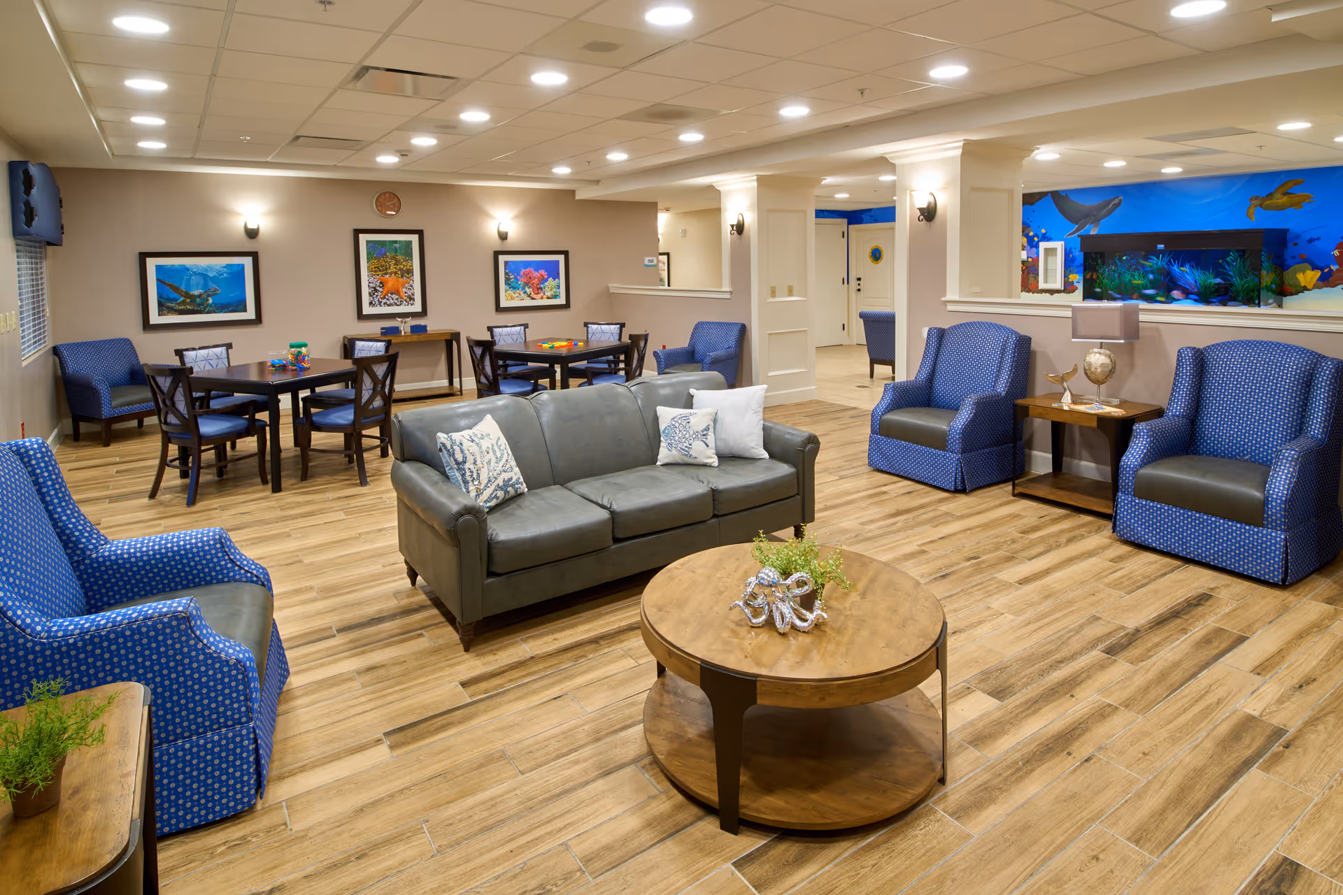 A spacious and well-lit common living area with wooden flooring, featuring a gray leather sofa with decorative pillows, three blue patterned armchairs, a round wooden coffee table with a small plant and decorative item, and two tables with chairs in the background. The walls are adorned with framed underwater-themed artwork, and there is a large aquarium built into a half wall. The ceiling has recessed lighting and a tiled design.