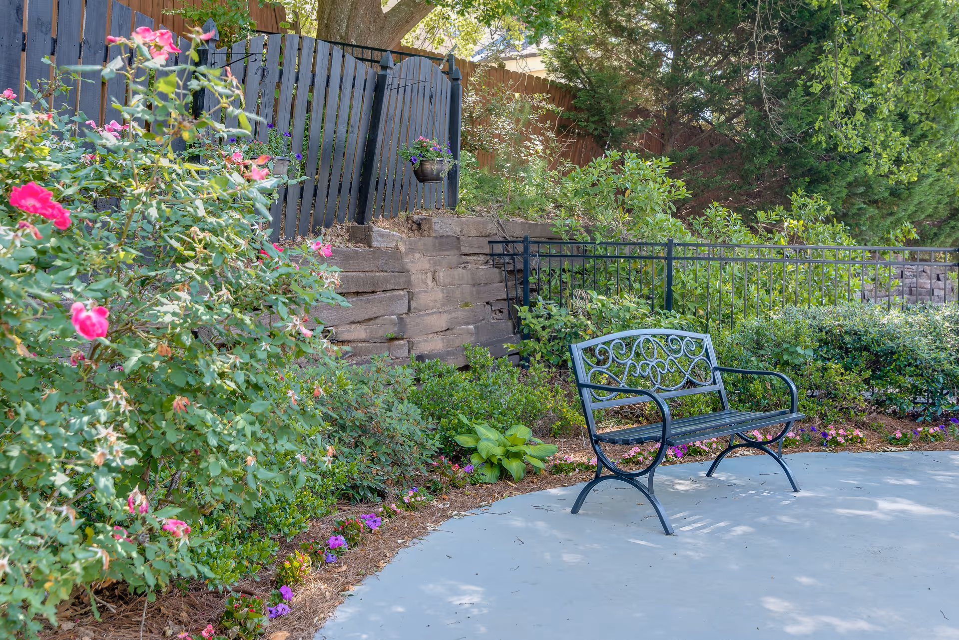 A black metal bench with decorative backrest sits on a concrete patio surrounded by lush greenery and flowering plants, including pink roses. Behind the bench is a black metal fence and a wooden fence with hanging flower pots, with trees and shrubs in the background.